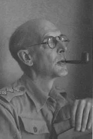 Older balding man in a military shirt smoking a pipe.