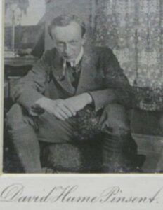 Man with an intense gaze leans forward, seated on an ottoman.