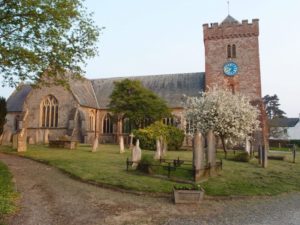 Modern photo of an old church of red and grey stone. There is a graveyard out front.