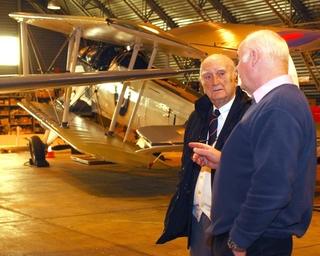 Two old men stand in front of a biplane.