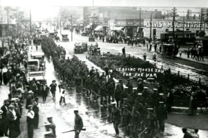 A black and white photograph of columns of Canadian soldiers lined up in the street, surrounded by watching crowds. 