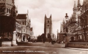 An old photograph of the streets of Plymouth. Ornate-looking buildings are on each side of the street. A church looms in the centre.