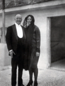 Janet and Frank stand outside an open doorway. Frank is in a tuxedo and Janet is in a dark outfit with black lace covering her hair.