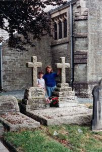Two children, a small boy and slightly taller girl, stand between two graves.