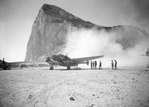Black and white photo of an old plane with its propellers spinning. It kicks up dust behind it. People stand nearby.
