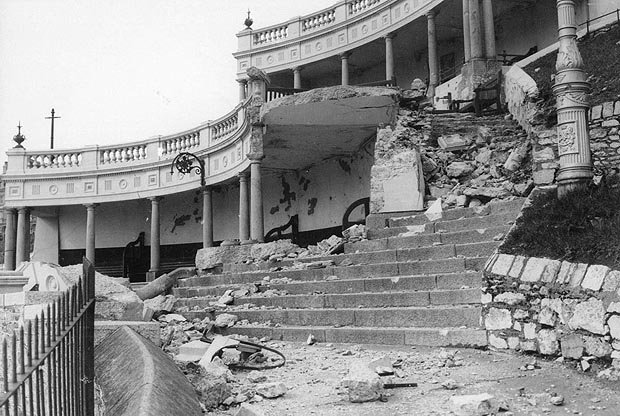 The partially destroyed remains of an ornate building with stone columns. Its staircase is now covered in rubble.