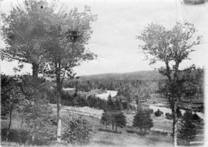 A faded black and white photograph of rolling hills, trees, and a river.