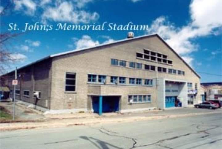 Modern photo of a large, simple building against a blue sky. It reads St John's Memorial Stadium.