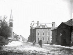 A damaged black and white photograph of a snowy open street, with a stone building in the background. Several people stand in the snow.