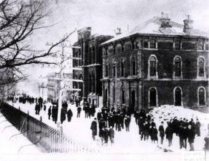 Black and white photograph. Dozens of people mill about outside the bank in the snow.