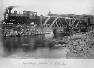 A faded black and white photograph of a train on a bridge. Men working the train, wearing dark clothes, stand in front and around the train.