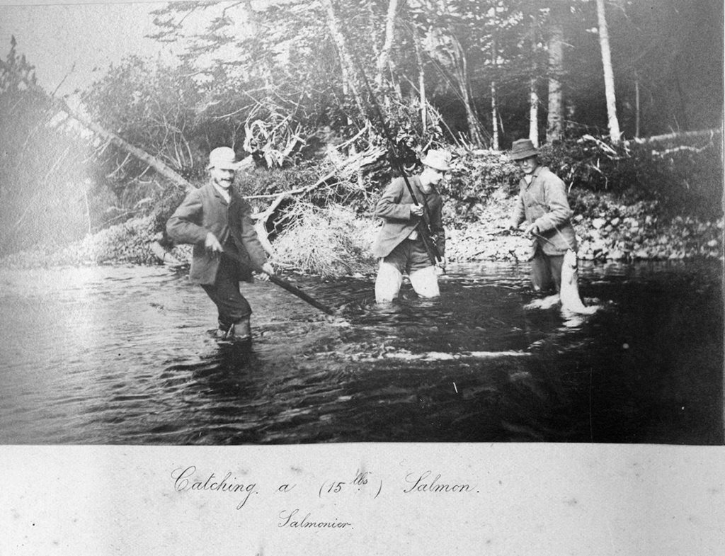 A black and white photograph of three men wading in a river, fishing. One man has snagged a 15 lb salmon.
