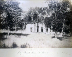 A faded black and white photograph of a small home surrounded by tall deciduous trees.
