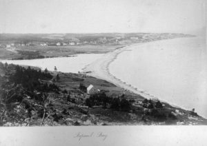 A faded black and white photograph of Topsail Bay, a curved stretch of land with scattered trees in the foreground and buildings in the distance. It is nestled against the water.