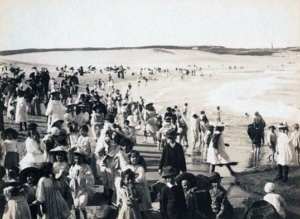 People in Victorian clothes throng in a black and white photo of a beach.