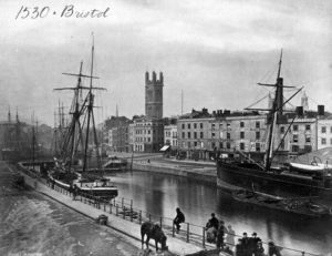 Black and white photograph of Bristol. A river or a canal, with buildings and a church on one side. Sail ships are anchored on either side.