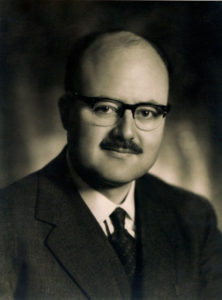 Headshot of a kindly-looking, balding man with a mustache.