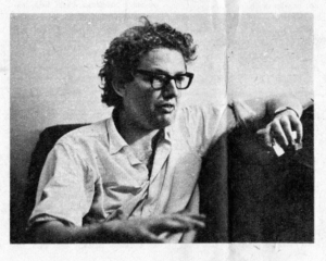 Black and white photograph of a white man with glasses and unkempt hair. He's moving his hands as he talks.