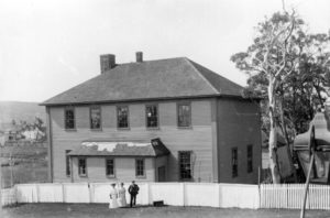 A simple two-story rectangular schoolhouse. Three people in Victorian garb stand outside.
