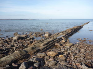 Modern photograph of an old pipe jutting out from a rocky beach and into the sea.