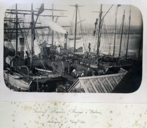 A faded black and white photograph of a dock surrounded by fishing ships. The dock is crowded with people.