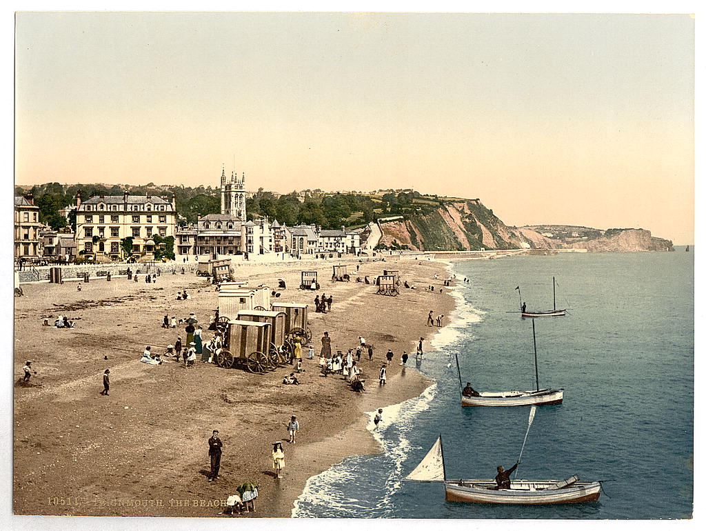 Old photograph of a beach spotted with people in Victorian clothes at the seaside. Small boats sit in the water.
