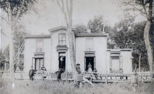 Black and white photograph of a white house behind a wood fence. Men and women in 1900s fashion stand in front of it.