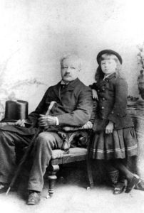 A faded black and white photograph of Robert John Pinsent, seated, as an older man. A young girl stands beside him.