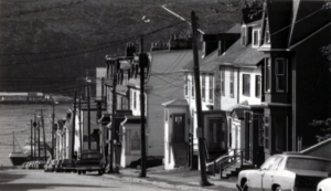 A black and white photograph of a comparatively modern street scene, with townhouses and telephone poles and a car.