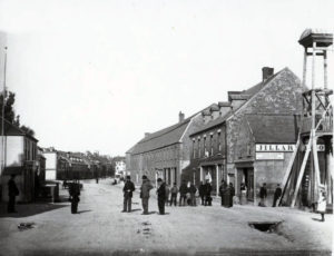 A black and white photo of a single street in Harbour Grace. People mill about outside.,