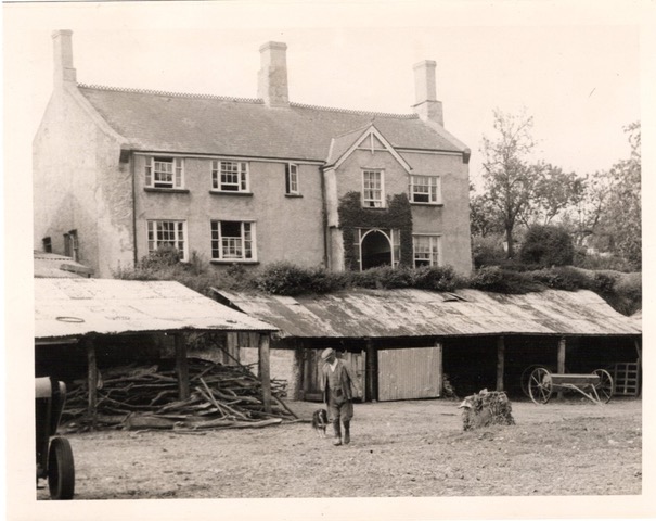 Black and white photograph of a simple multi-storey building fronted by sloped-roof farm structures.