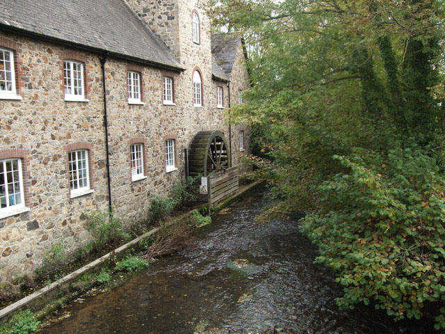 A sand-coloured stone building alongside a small river. An old wooden waterwheel is built into its side.