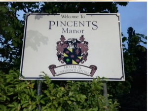 Modern photograph of a sign surrounded by trees. It reads Welcome to Pincents Manor Hotel & Conference Center. It features a crest in yellow and black, with a red lion, three red crowns, and a green tree.