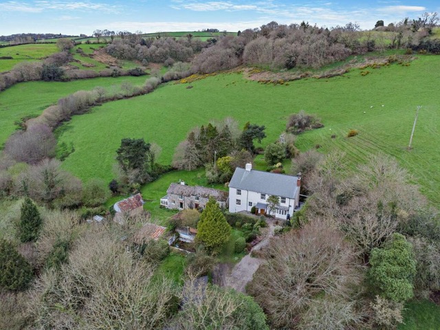 A modern house sits next to an older structure. It is surrounded by green fields and trees.